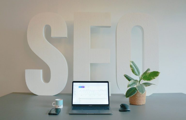 A workspace featuring a laptop, coffee cup, plant, and large "SEO" letters in the background.